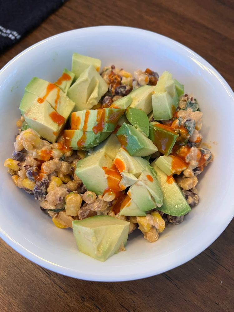 A white bowl holding a salad with quinoa, corn, black beans, and a creamy chipotle dressing top with diced avocado