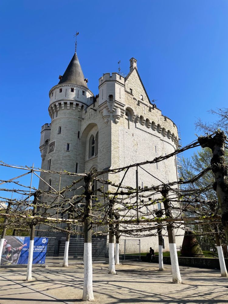 A white stone 'castle' with a tall, pointed tower and battlements stands against a clear blue sky. In the foreground, a trellis structure with bare vines is visible