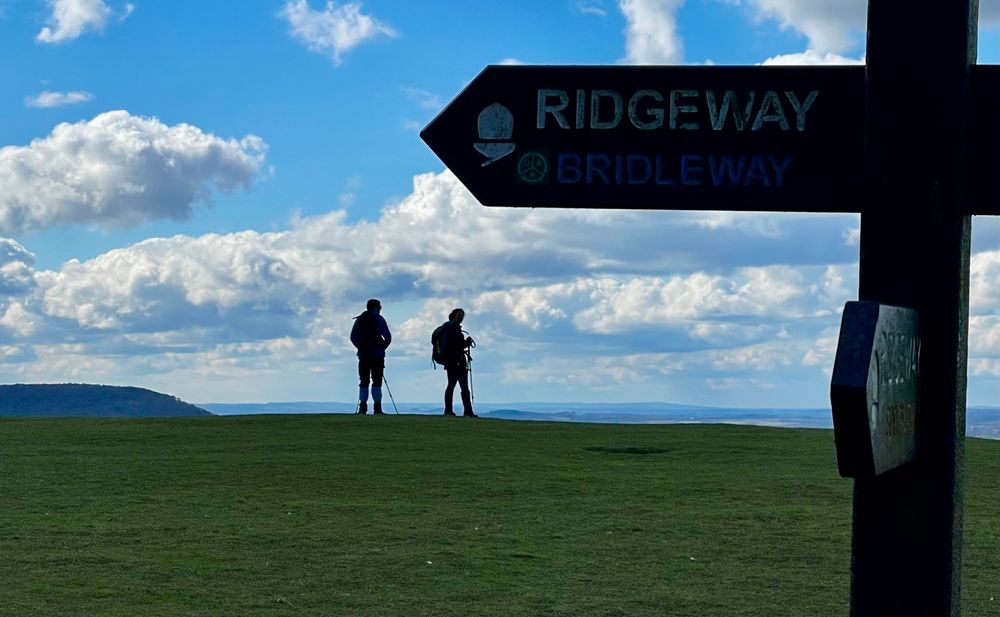 The image shows two walkers on Whiteleaf Hill above Princes Risborough looking at the view with a Ridgeway National Trail sign in the foreground.