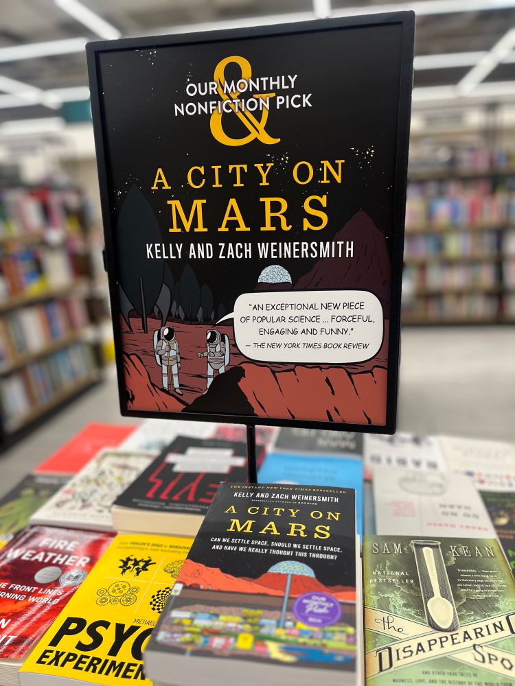 A tabletop book display in a Barnes & Noble, with a pile of books on it and a sign reading “Our Monthly Nonfiction Pick: A City on Mars, by Kelly & Zach Weinersmith”