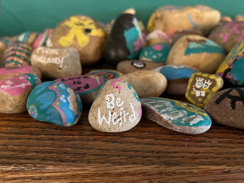 Closeup of a bunch of painted stones on a wooden shelf. One stone says “Be Weird” in white letters.