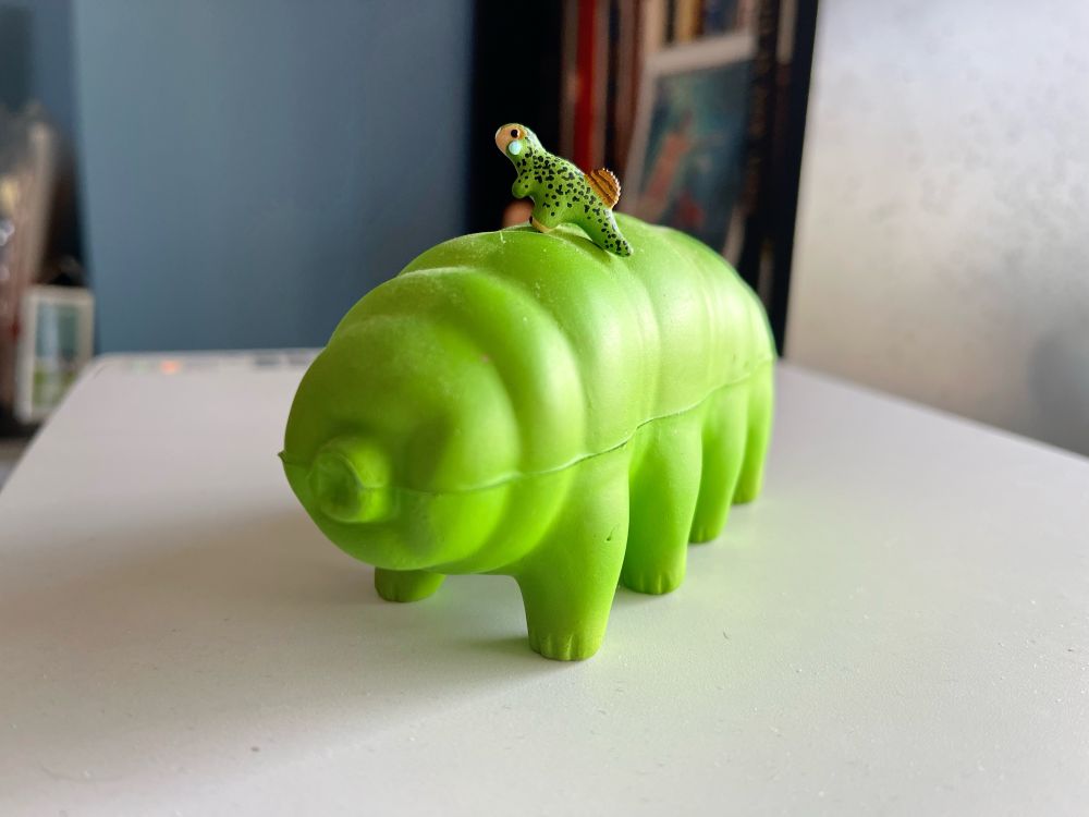 A bright-green foam tardigrade stands on top of a printer, with a very, very small painted clay model of a green, black-spotted psittacosaur on its back.