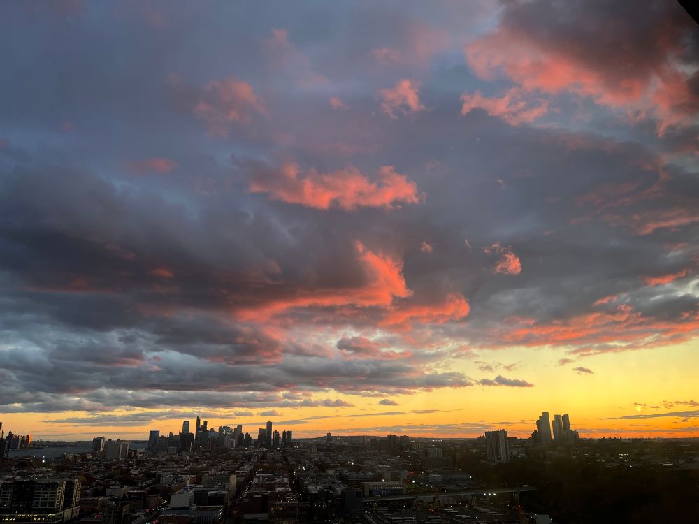 High-angle view of pink and gray sunset clouds over a city, with yellow and orange light over the horizon.