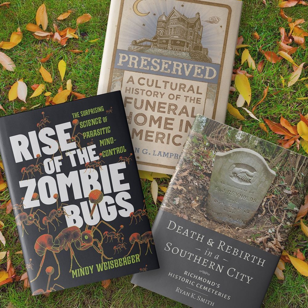 An overhead view of three hardcover books on grass covered with colorful fall leaves. The books are "Rise of the Zombie Bugs," "Preserved: A Cultural History of the Funeral Home in America," and "Death and Rebirth in a Southern City"