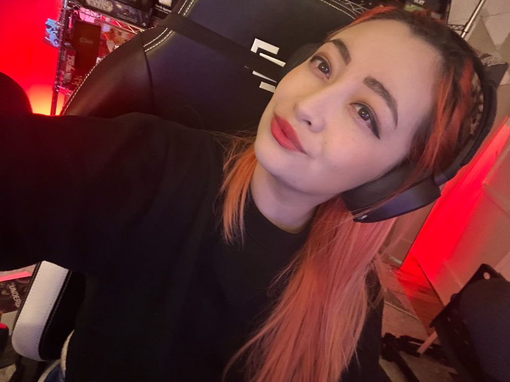 An orange haired girl in a black sweater sitting in a chair with a headset on. 