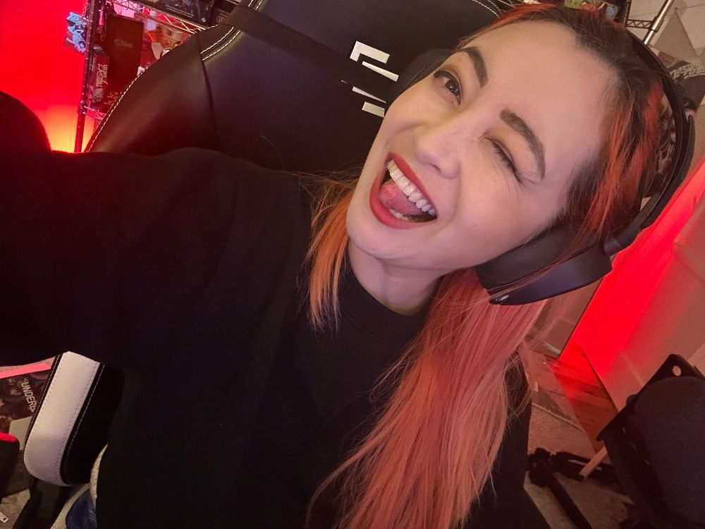 An orange haired girl with a black headset on. She is wearing a black sweater and is winking.