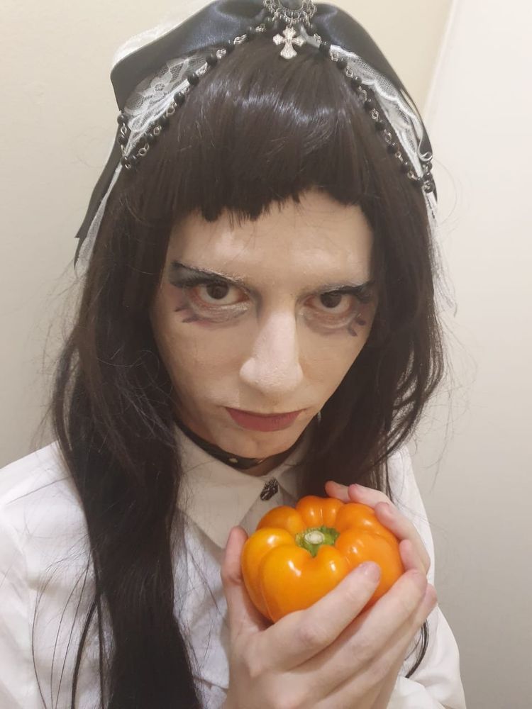 Uma pessoa com peruca preta e maquiagem gótica segurando um pimentão em formato de uma mini abóbora. 
