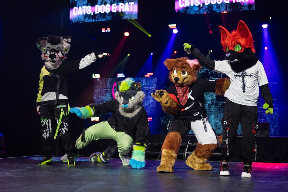 JD Puppy, a brown floppy eared fursuiter, performs with Zero, Kito, and Shayde, the other fursuit performers in the group Cats, Dog & Rat at Furry Weekend Atlanta. They are wearing techgear in neon green, black, white, and red clothing. Zero and Shayde are standing on the outside, raising arms to frame the roof of a house. Kito and JD are squatted on the inside, as if peeking through the house. One of JD's ears is unflopped.