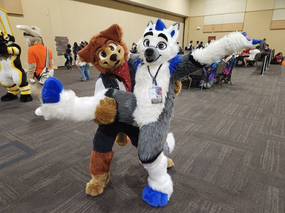 JD Puppy,  a brown floppy eared dog fursuiter, holds the lifted leg of Bexis, a white and gray canine suiter with royal blue highlights for hair, eartips, fingers, and toes,  in an open convention center expo hall area