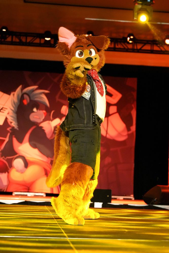 JD Puppy, a brown floppy eared fursuiter, performs on the Anthro Weekend Utah stage highlighted by yellow stage lights. He is wearing his typical black denim vest, white t-shirt, red bandana, and black cargo shorts. He is pointing at the camera in the middle of a dance routine. One of JD's ears is unflopped.