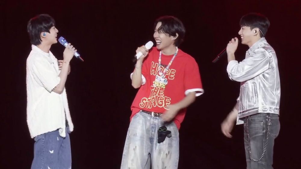A photo of Jin, Jhope and Jungkook of BTS on stage holding mics while laughing.