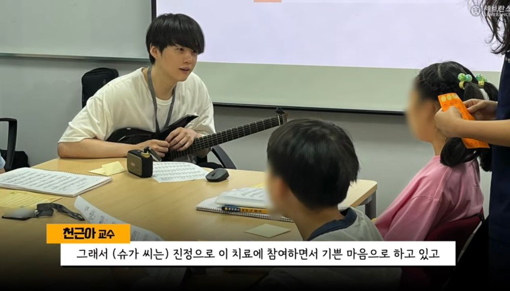 Suga sitting on a chair playing guitar to kids