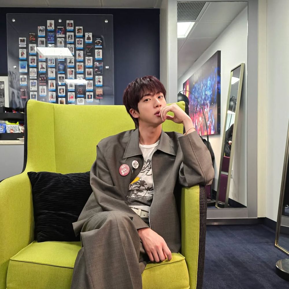 Jin of BTS sitting on a light yellow-green sofa, wearing a light grey jacket and pants and a white graphic t-shirt underneath. With one hand on his lap, the other hand on his chin.