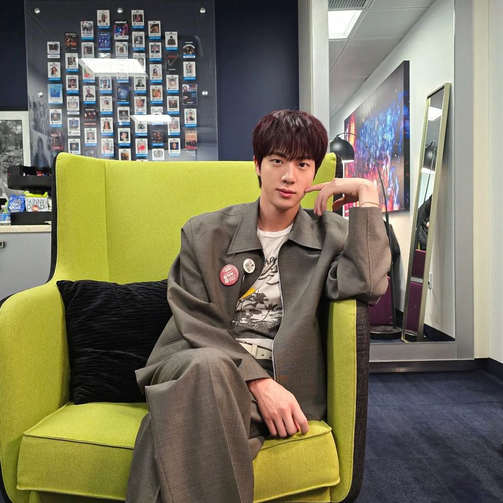 Jin of BTS sitting on a light yellow-green sofa, wearing a light grey jacket and pants and a white graphic t-shirt underneath. Posing with one hand on his lap, the other hand poking his cheeks.