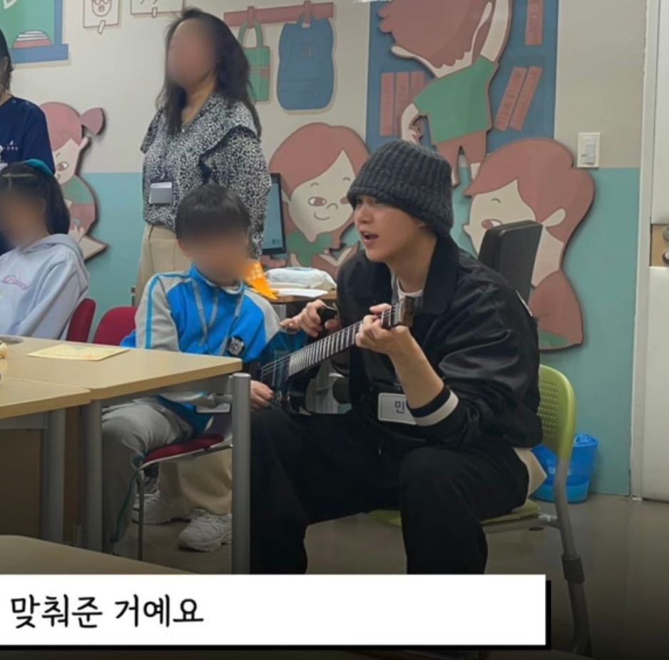 Suga sitting on a chair playing guitar to kids 