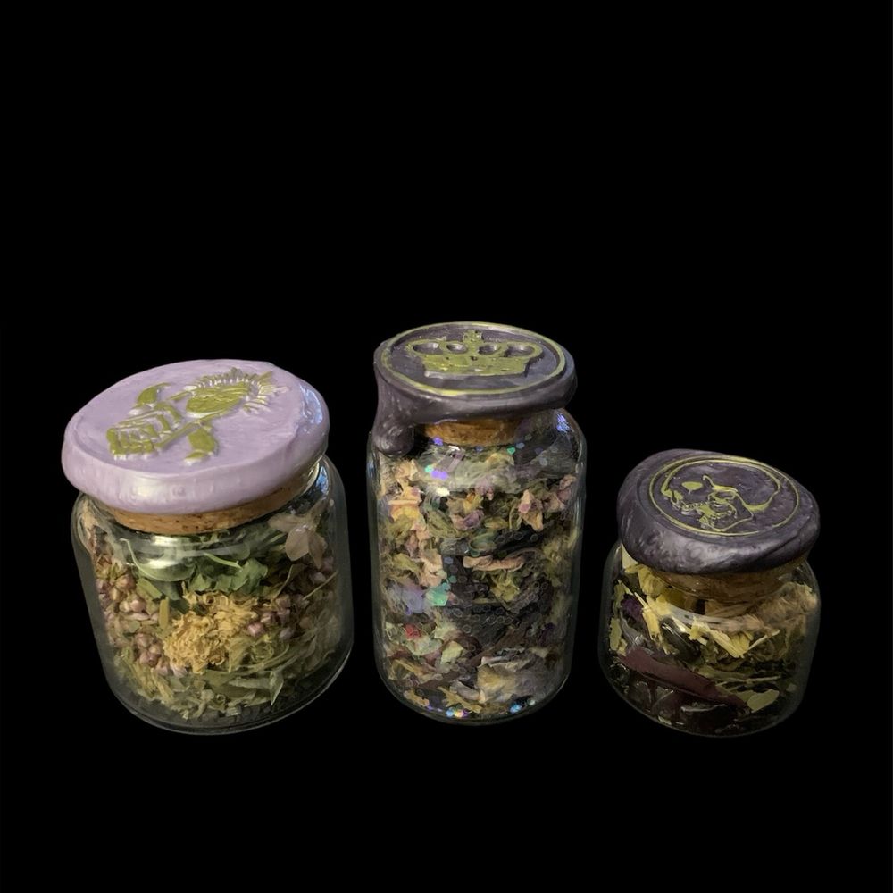 Three potions bottles on a black background. The first is wide and sealed with light purple wax and a gold skeleton hand stamp, and contains various herbs and flowers; the second is taller and is sealed with dark purple wax and a gold crown stamp, and contains various herbs and flowers as well as glitter; the third is short and sealed with dark purple wax and a gold skull stamp, and filled with flowers and herbs as well.