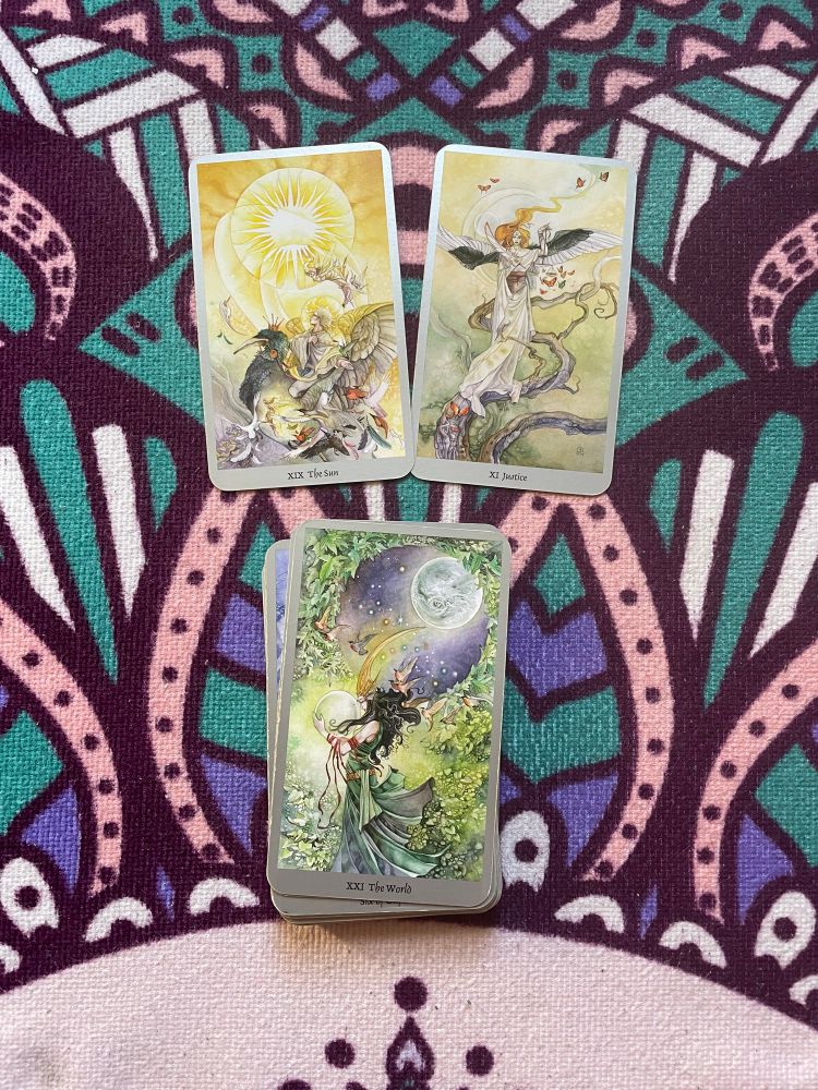 three tarot cards on a colorful background. On the top left is the sun. A man illuminated by Gold rides the bird.. on the top right is justice. An angel holds a set of scales and stands on a tree branch. Below is the world card. A woman looks lovingly at a small world. She’s holding her hands while through a window. We can see another world in the background floating in space.