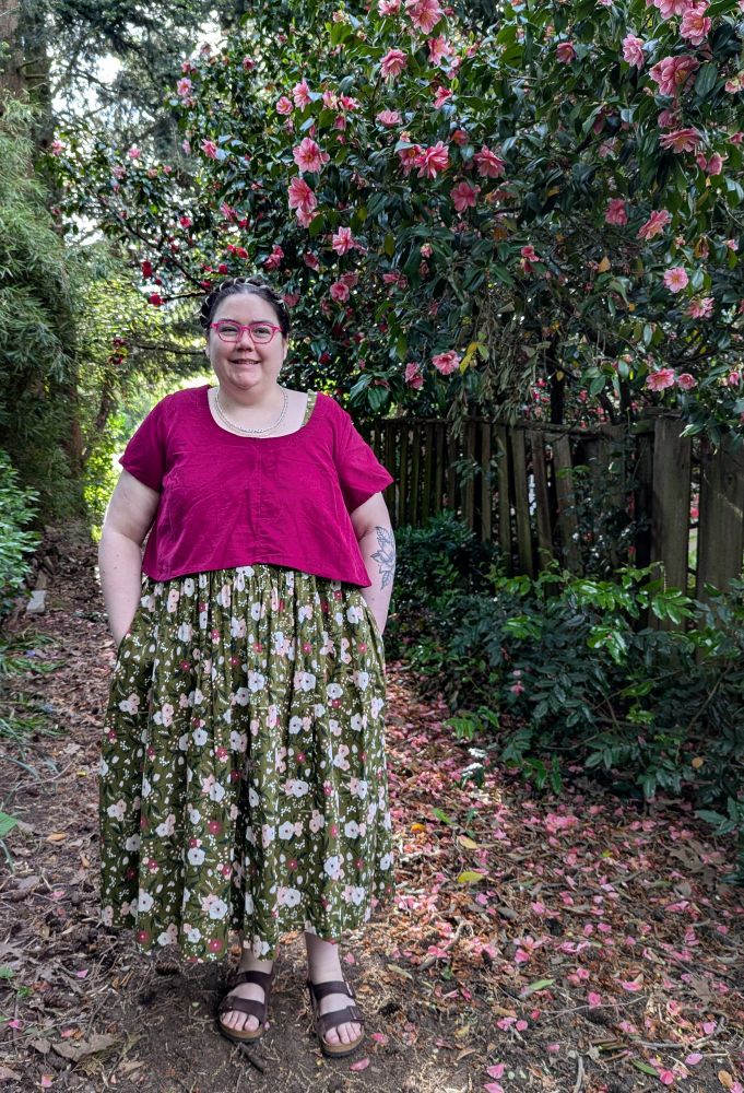 A fat person standing in a garden, wearing a floral print dress and a corduroy top.
