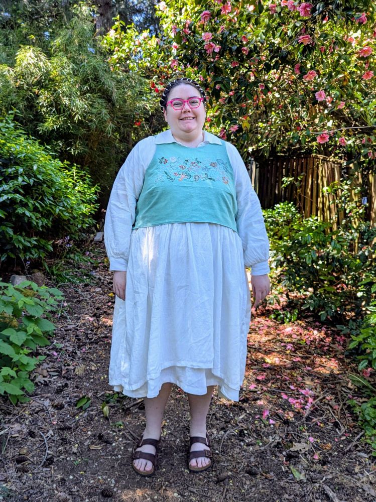 A fat white person standing in a garden, wearing a white dress, an embroidered vest, and sandals.