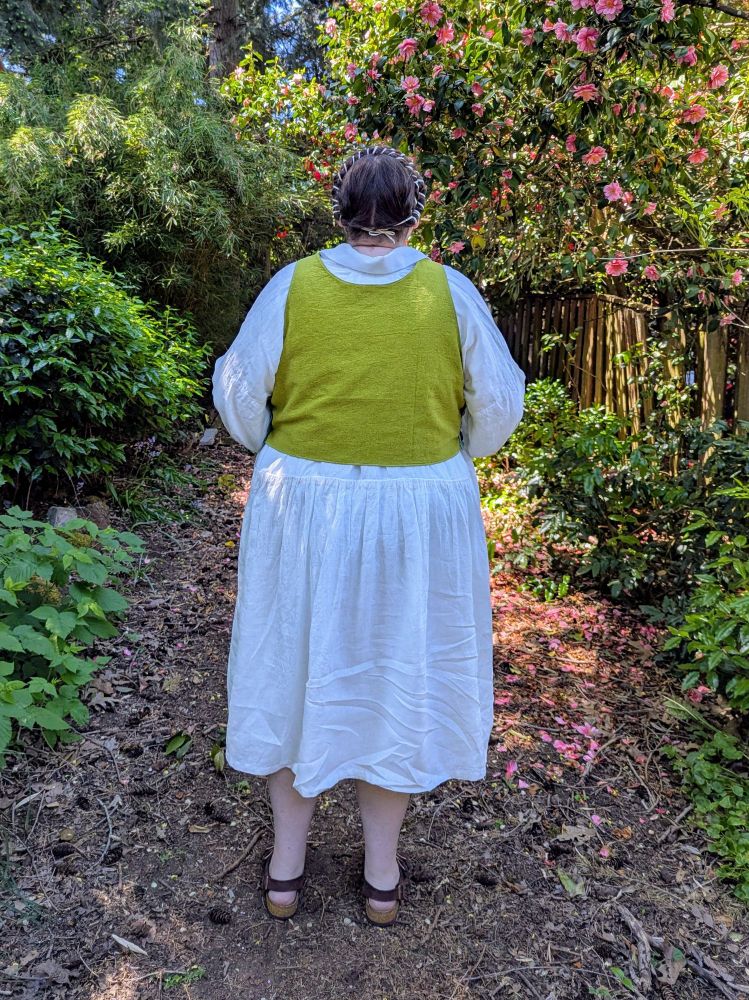 A fat white person standing in a garden, wearing a white dress, an embroidered vest, and sandals. They are turned away from the camera, highlighting the contrasting back panel.