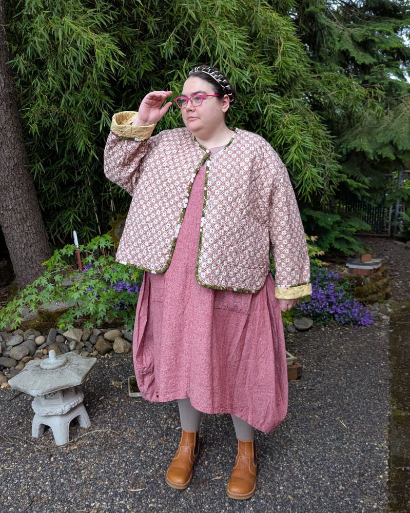 A fat white person standing in a garden, wearing a long dress, quilted jacket, and boots.
