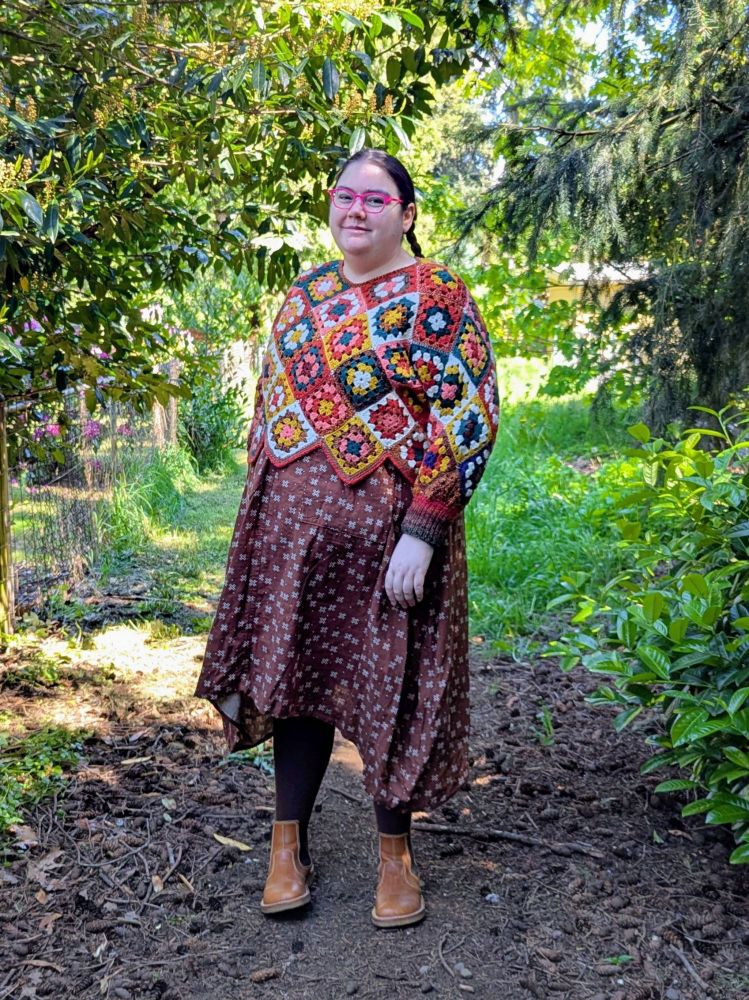 A fat white person standing in a garden, wearing a brown dress with an overall plus pattern, a colorful crocheted sweater, and boots.