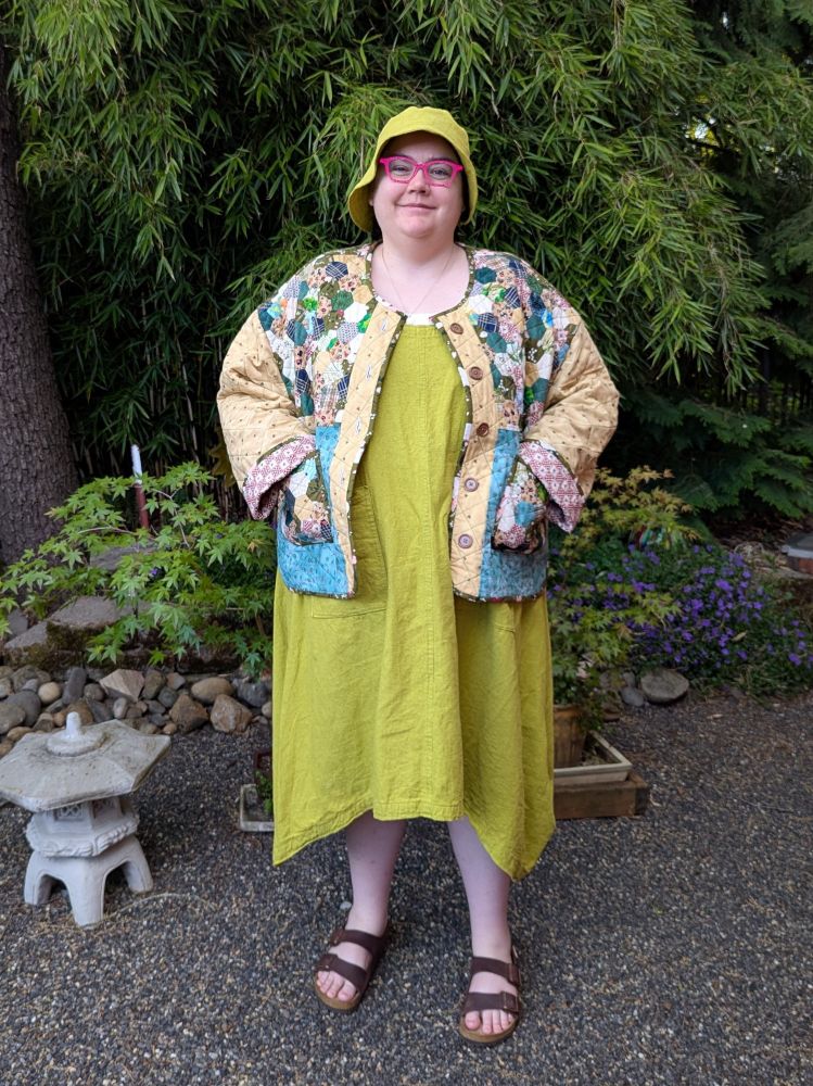 A fat person standing in a garden wearing a long dress with matching bucket hat, a quilted, patchwork jacket, and sandals.