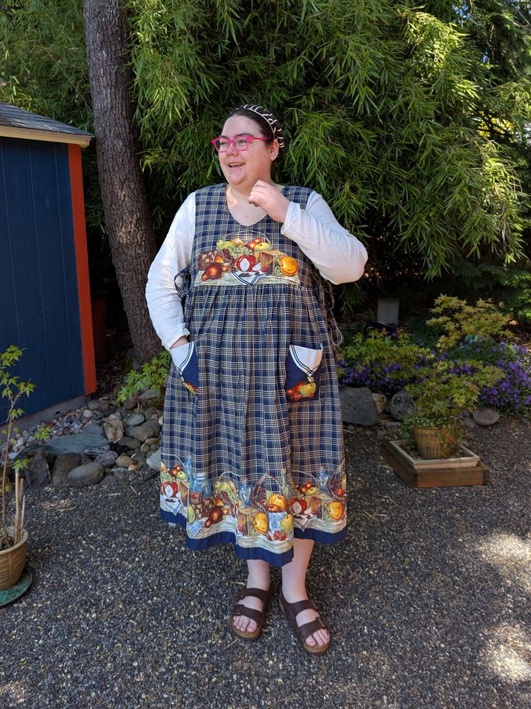 A fat white person standing in a garden, wearing a navy dress with pictures of a picnic on it, a white shirt, and sandals. They are looking away and touching their necklace.