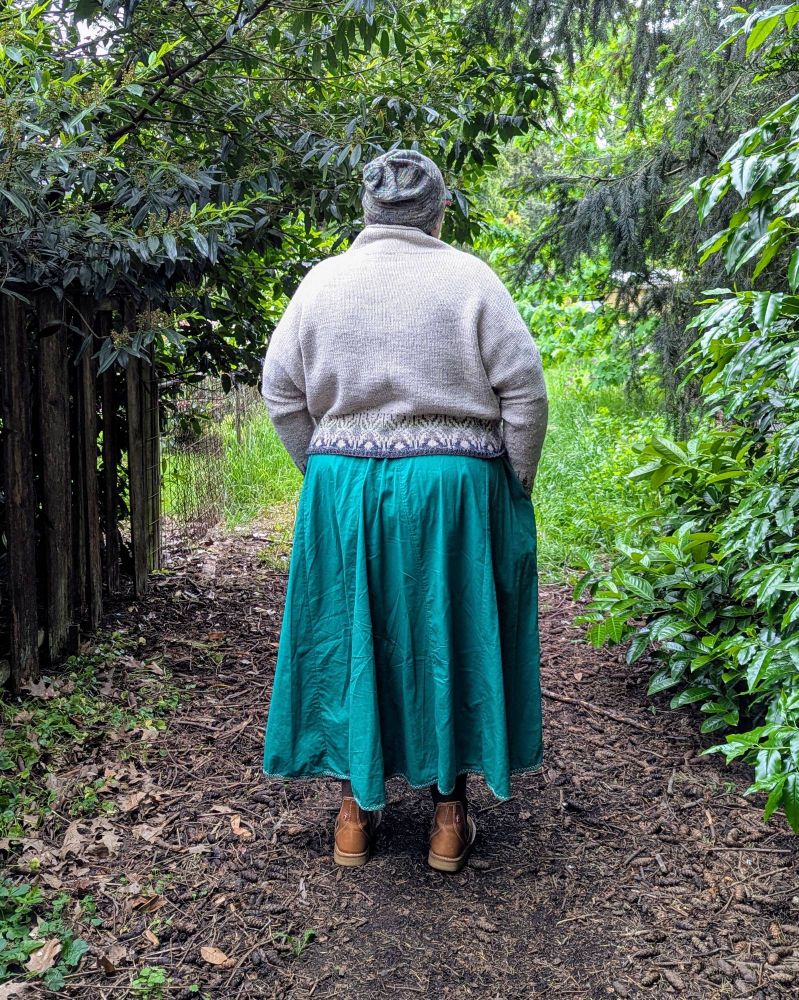 A fat white person standing in a garden, wearing a long dress, hand-knit sweater and hat, and boots. They are turned away from the camera.