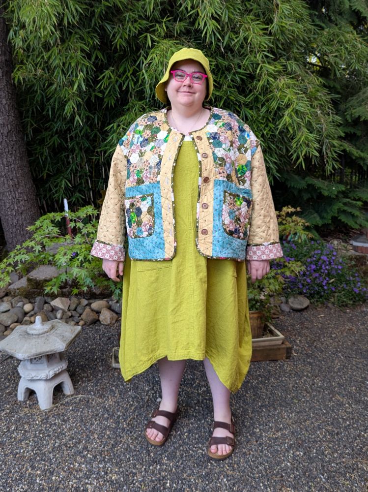 A fat person standing in a garden wearing a long dress with matching bucket hat, a quilted, patchwork jacket, and sandals.