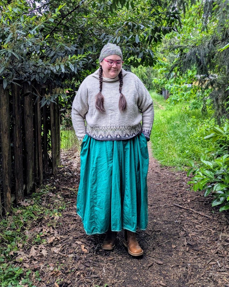 A fat white person standing in a garden, wearing a long dress, hand-knit sweater and hat, and boots.