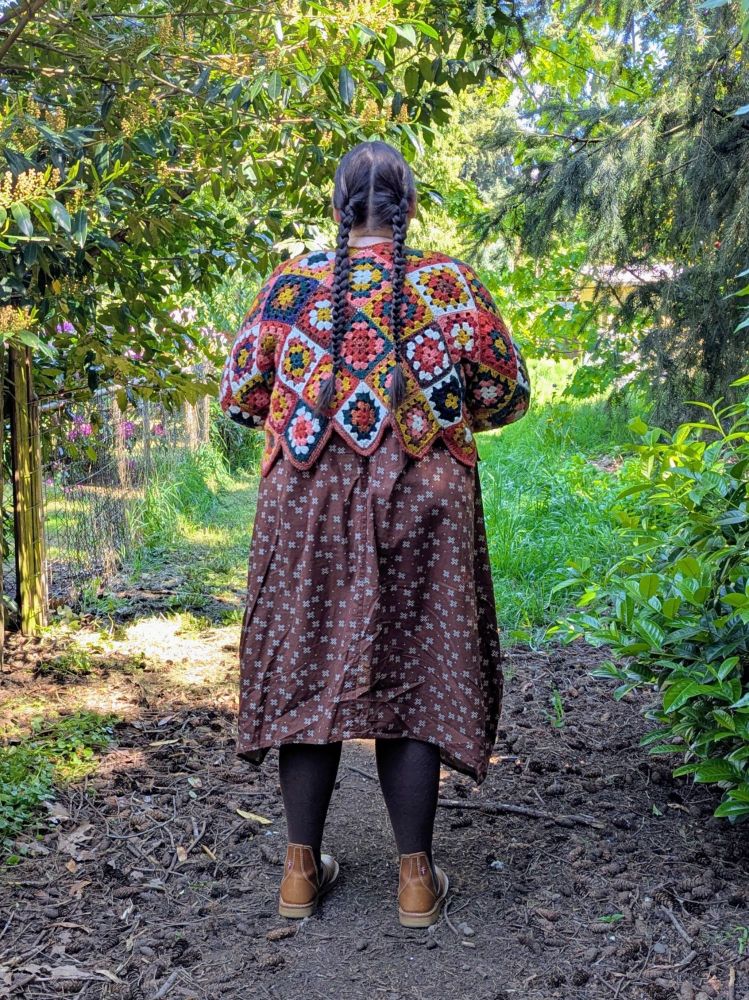 A fat white person standing in a garden, wearing a brown dress with an overall plus pattern, a colorful crocheted sweater, and boots. They are turned away from the camera, showing their two long braids.