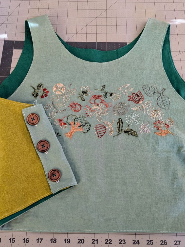 A cropped flat-lay of the embroidered vest, revealing the darker lining, button placket, and embroidery. The embroidery motifs include trees, leaves, flowers, birds, and insects.