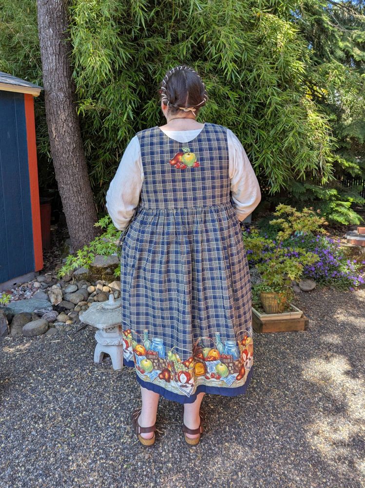 A fat white person standing in a garden, wearing a navy dress with pictures of a picnic on it, a white shirt, and sandals. They are turned away from the camera, showing the fruit print on the back of the dress bodice.