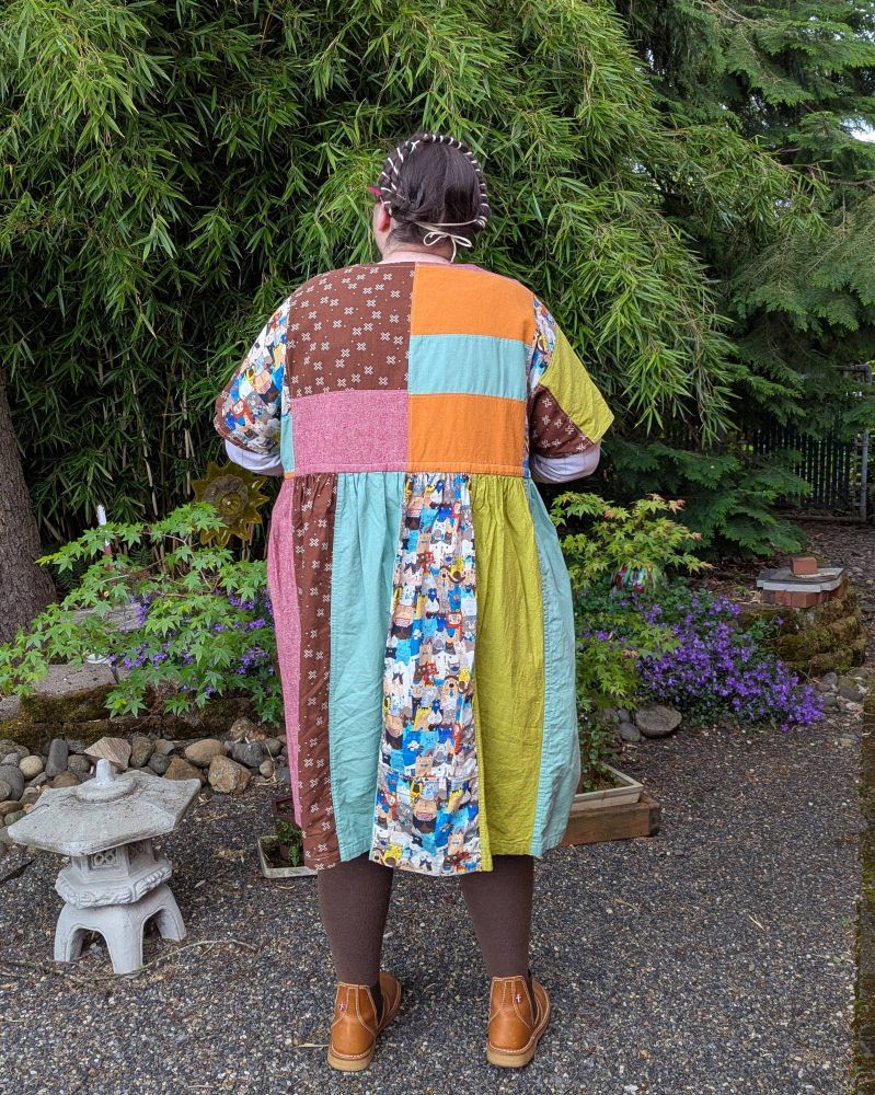 A fat white person standing in a garden, wearing a colorful patchwork dress, a long-sleeved shirt, and boots. They are turned away from the camera, showcasing the patchwork fabric on the back of the dress.