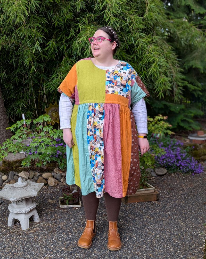 A fat white person standing in a garden, wearing a colorful patchwork dress, a long-sleeved shirt, and boots.