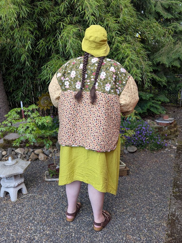 A fat person standing in a garden wearing a long dress with matching bucket hat, a quilted, patchwork jacket, and sandals. They are turned away from the camera, highlighting the back of the jacket.