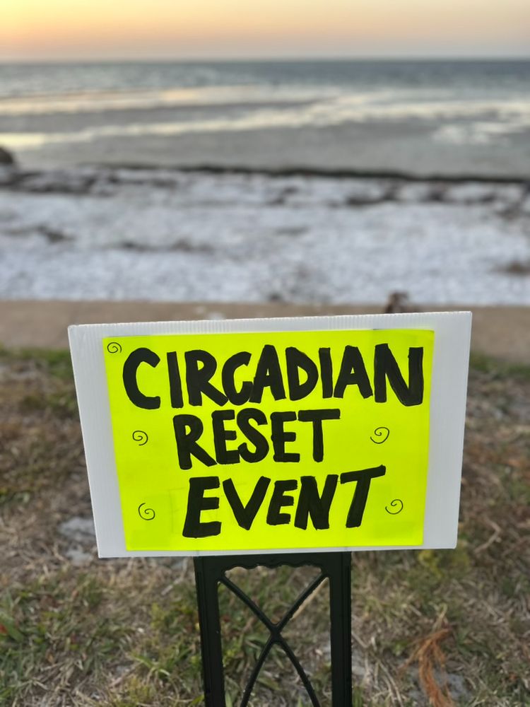 Picture of a neon yellow sign with black block letters stating CIRCADIAN RESET EVENT. A blurry background shows water from Tampa Bay.