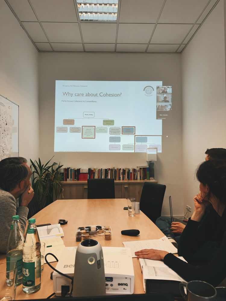 Die Aufnahme zeigt eine kleine Gruppe von Personen, die an einem Konferenztisch sitzen und einer Präsentation folgen. An der Wand wird eine PowerPoint-Folie projiziert, die sich mit dem Thema “Party Group Cohesion in Committees” befasst. 