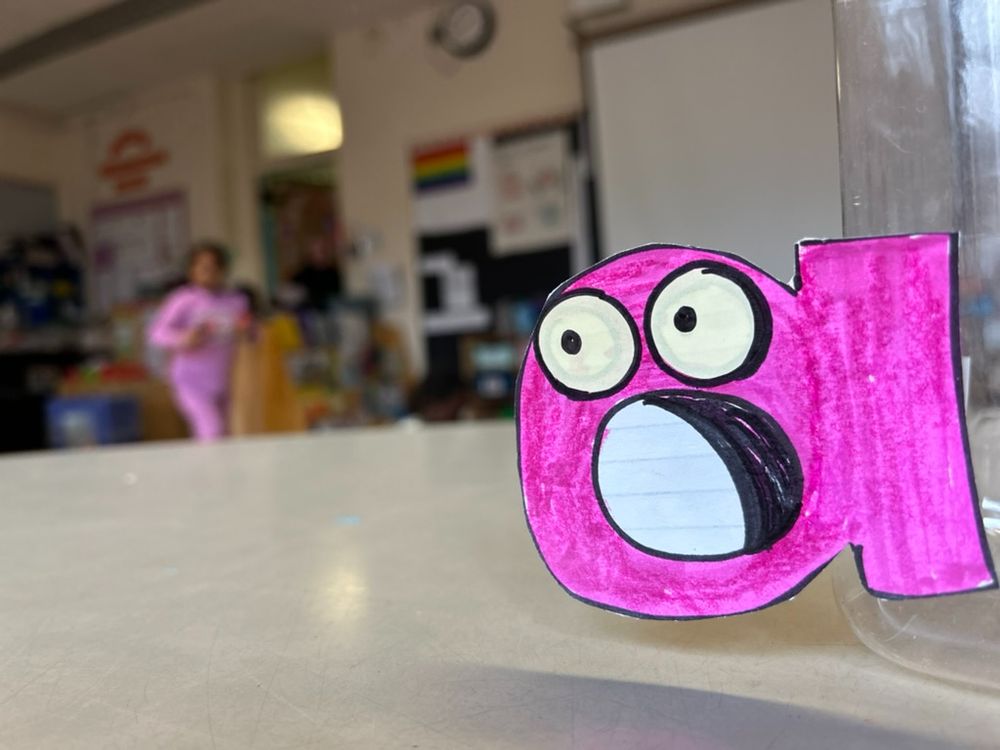 There's a pink lowercase a that looks like it's screaming. It's a close up and the classroom is blurry. It's a kindergarten classroom. 
I drew the a with big eyes and a screamy mouth.