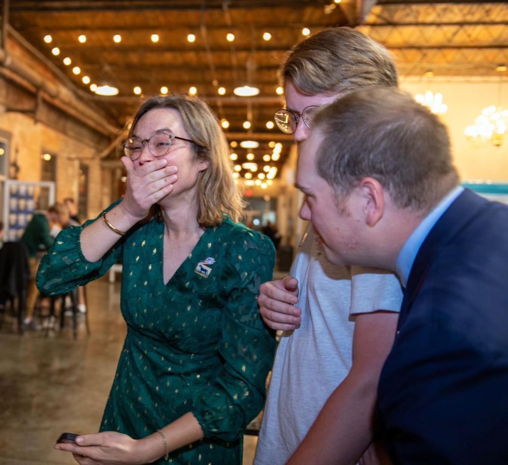 Emma covers her mouth, crying after receiving the news that she won. She’s standing next to two white men looking over her shoulder at the election results. 