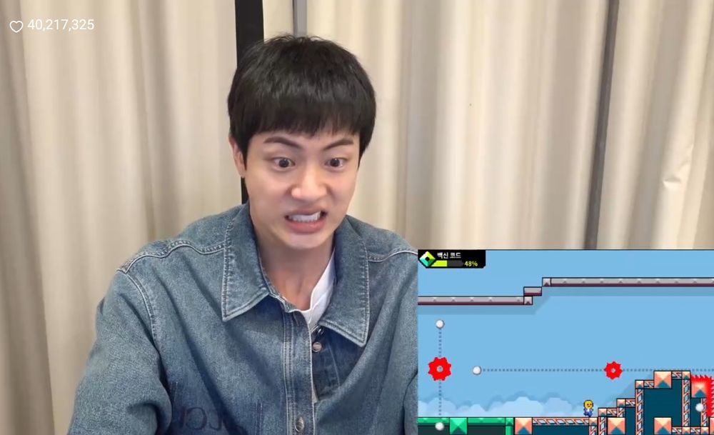 BTS' Jin playing computer games on Weverse live. He is wearing a Gucci jean t-shirt and the computer screen is showing in the lower, right corner. Jin is grimacing with frustration because he lost in the game.