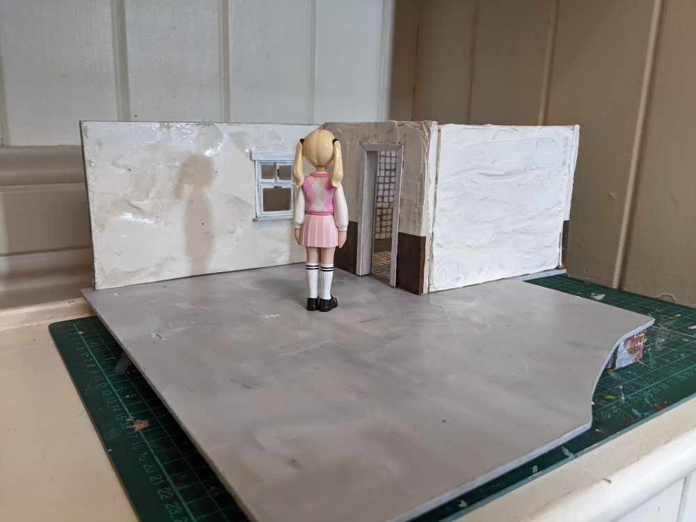 A solitary pink uniformed Pop stands with its back turned, in the middle of a bare miniature room walls now partially erected.