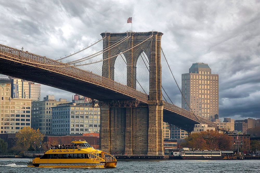 A bright yellow New York Water Taxi passing under the Brooklyn Bridge.