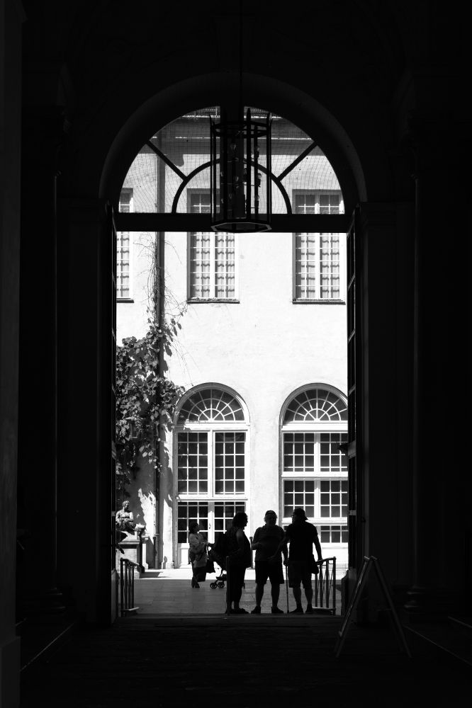 Black and white showing a big door in front of a light wall of another house with big windows and three people as silhouettes. 
