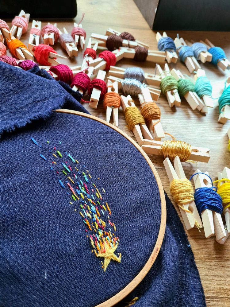 An embroidered falling star/comet with colourful yarns on dark blue cloth. In the background there are clothesline clips with lots of different yarns rolled up on them. 