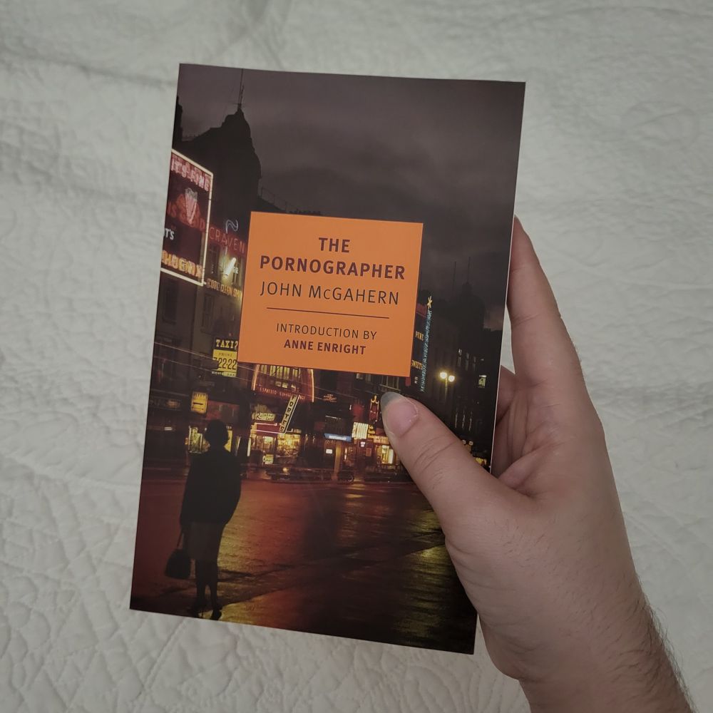 Picture of me holding the book The Pornographer by John McGahern, published by New York Review of Books. The book has its signature square in the middle in a bright orange color with dark red lettering for the title. The cover image features a photograph of an illuminated street at night, and the figure of a woman at the edge of the road. 