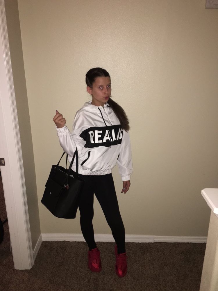 Famous meme of a girl posing and pursing her lips sassily in her hallway. She wears a white hoodie with the word "REALIST" on it, black leggings, red shoes, and holding a large black purse in her right arm. 