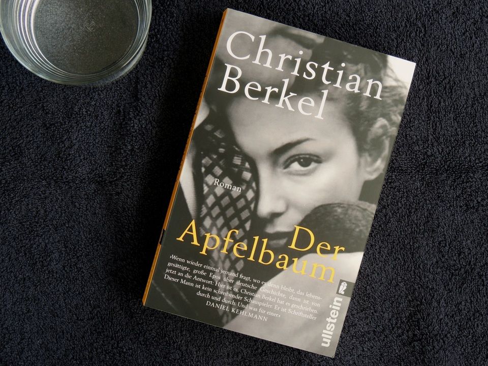 Christian Berkels Roman "Der Apfelbaum" liegt auf einem schwarzen Strandtuch. In der linken oberen Ecke steht ein Glas Wasser.