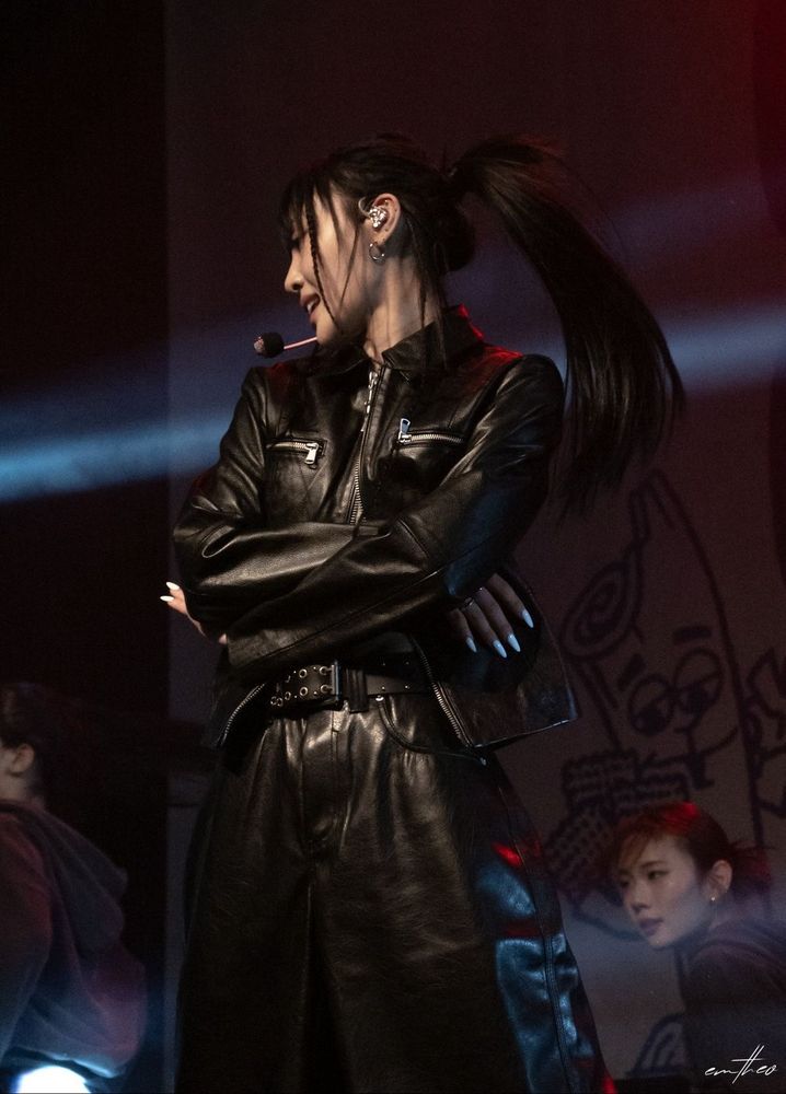 Photo of yves in a black leather jacket and pants performance outfit with her arms crossed and her long black ponytail whipping around as she faces to the side 